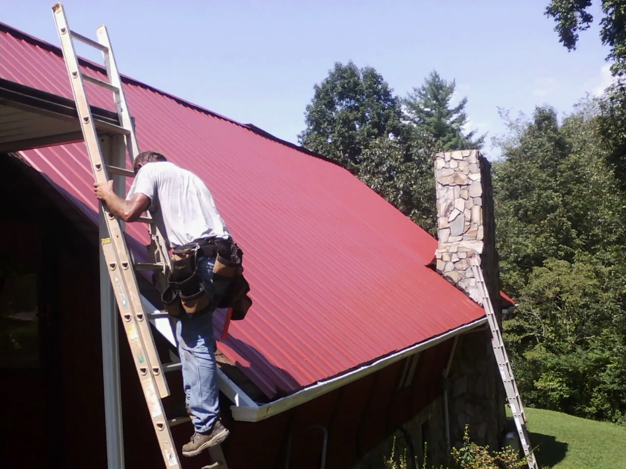 Roofer on a ladder beside a red metal roof and stone chimney — Soffit & Fascia Repair by Roofing Brothers in Turners Falls